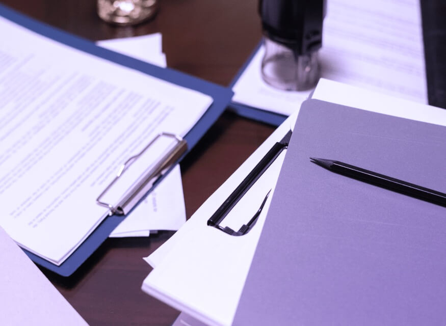 Na biurku znajdują się dokumenty biznesowe, teczki, clipboard z umową oraz długopis przygotowany do podpisu. Obok leżą okulary, co podkreśla profesjonalny i formalny charakter miejsca pracy. 